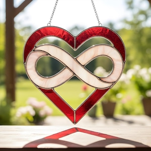 Könnte beinhalten: Ein herzförmiger Glas-Suncatcher mit rotem Rand und einem weißen Unendlichkeitszeichen in der Mitte. Er hängt an einer silbernen Kette. Der Hintergrund ist eine verschwommene Szene im Freien. Die Breite beträgt ca. 15 cm.