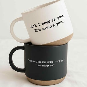 May include: Two ceramic mugs, one white and one black, stacked on top of each other. The white mug has the text "All I need is you. It's always you." The black mug has text in Russian.