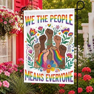 May include: A white garden flag with the text "WE THE PEOPLE MEANS EVERYONE" in rainbow colours. The flag features illustrations of raised fists in various skin tones, a rainbow, and floral designs. The flag is displayed outdoors in a garden setting.