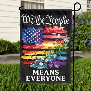 May include: A black garden flag with the text "We the People MEANS EVERYONE." The flag features a colorful watercolor design of the American flag with rainbow stripes and floral accents. The flag is hanging on a black metal stand.