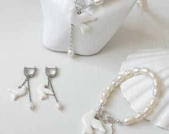 Puede incluir: Un conjunto de joyas con un collar de perlas, una pulsera y pendientes. Cada pieza tiene un colgante de paloma blanca y detalles de metal plateado. Las perlas son de color blanco roto y de forma ovalada.