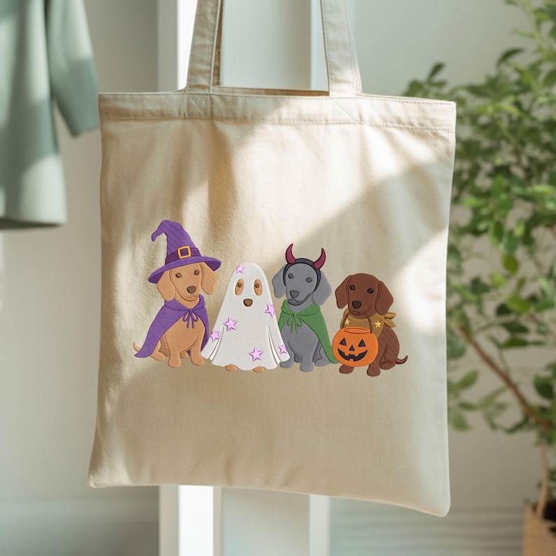 Puede incluir: Una bolsa de tela beige con un estampado gr&aacute;fico de cuatro perros salchicha disfrazados de Halloween. Un perro lleva un sombrero de bruja y capa morados, otro es un fantasma con estrellas rosas, otro tiene cuernos de diablo y capa verde, y otro sostiene una calabaza.