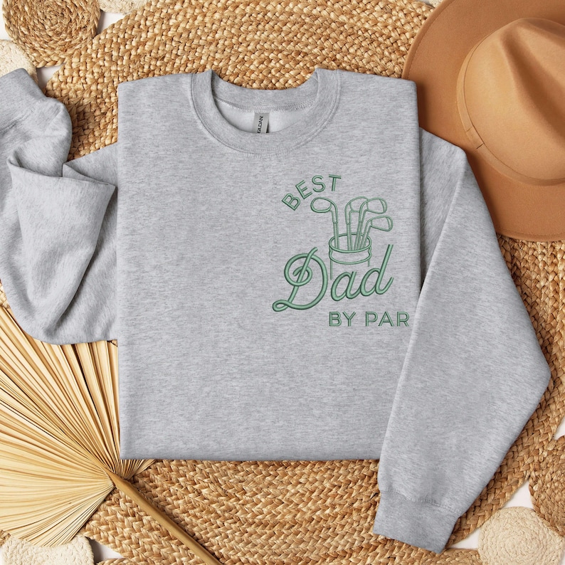 May include: A light gray sweatshirt with the text "BEST Dad BY PAR" in green. The design includes a golf club graphic. The sweatshirt is displayed on a woven surface with a brown hat in the background.