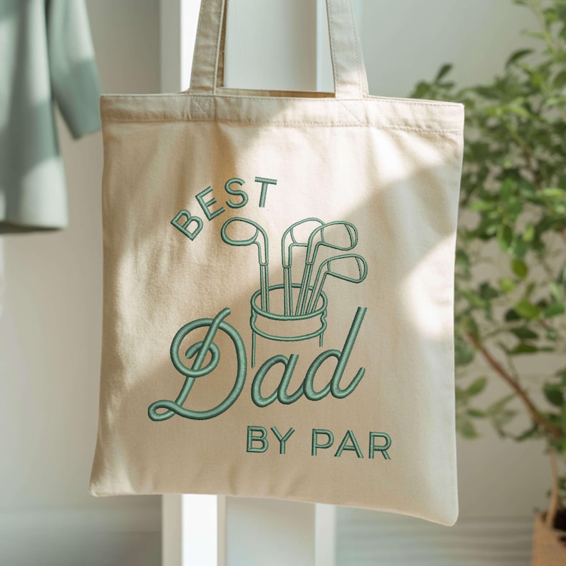 May include: Cream-colored canvas tote bag with green embroidered text and golf club graphic. The text reads "BEST Dad BY PAR". The bag has two handles and is likely made of cotton or a similar fabric.