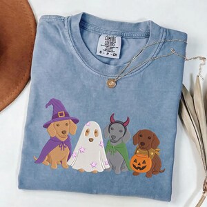 Puede incluir: Una camiseta azul de cuello redondo con un estampado gr&aacute;fico de cuatro perros con disfraces de Halloween. Un perro lleva un sombrero de bruja y una capa morada, otro es un fantasma con estrellas rosas, otro tiene cuernos de diablo rojos y capa verde.