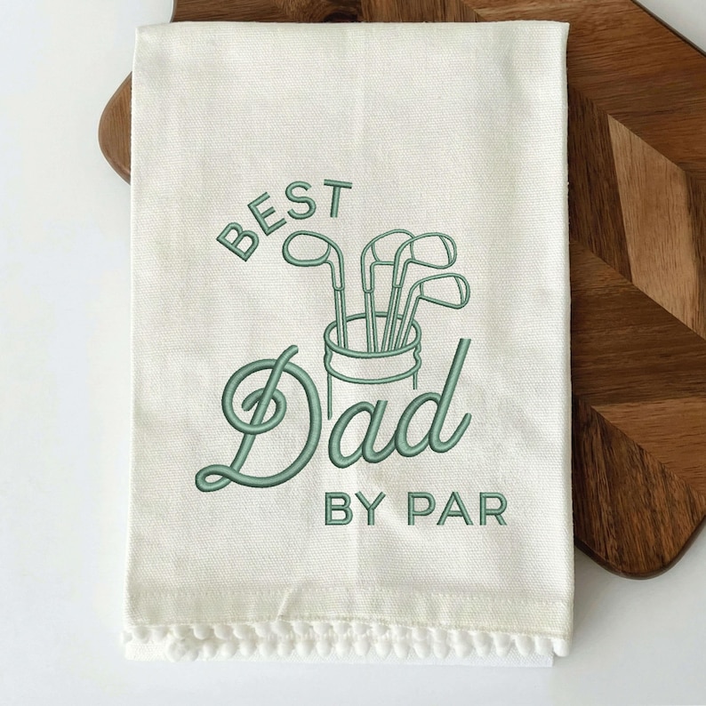May include: Off-white golf towel with green embroidered text and golf club graphic. The text reads "BEST Dad BY PAR". The towel has a fringe trim along the bottom edge. The towel is displayed on a wooden cutting board.