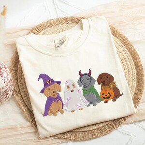 Puede incluir: Una camiseta de color crema con un gr&aacute;fico bordado de cuatro perros salchicha con disfraces de Halloween: uno de brujo con sombrero y capa morados, uno de fantasma con estrellas rosas, uno de diablo con cuernos rojos y capa verde, y otro con una calabaza.
