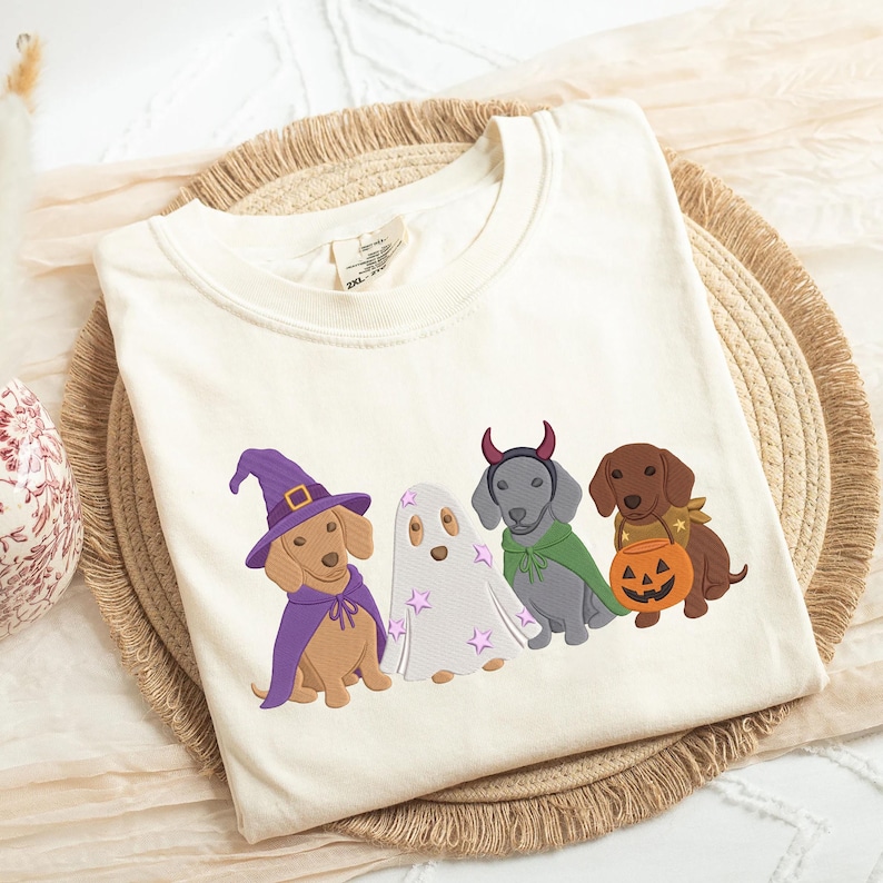 Puede incluir: Una camiseta de color crema con un gr&aacute;fico bordado de cuatro perros salchicha con disfraces de Halloween: uno de brujo con sombrero y capa morados, uno de fantasma con estrellas rosas, uno de diablo con cuernos rojos y capa verde, y otro con una calabaza.