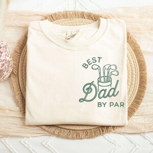 May include: Cream-colored t-shirt with the text "BEST Dad BY PAR" in green script and a golf club graphic. The shirt is folded and resting on a woven mat.
