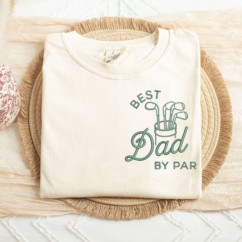 May include: Cream-colored t-shirt with the text "BEST Dad BY PAR" in green script and a golf club graphic. The shirt is folded and resting on a woven mat.
