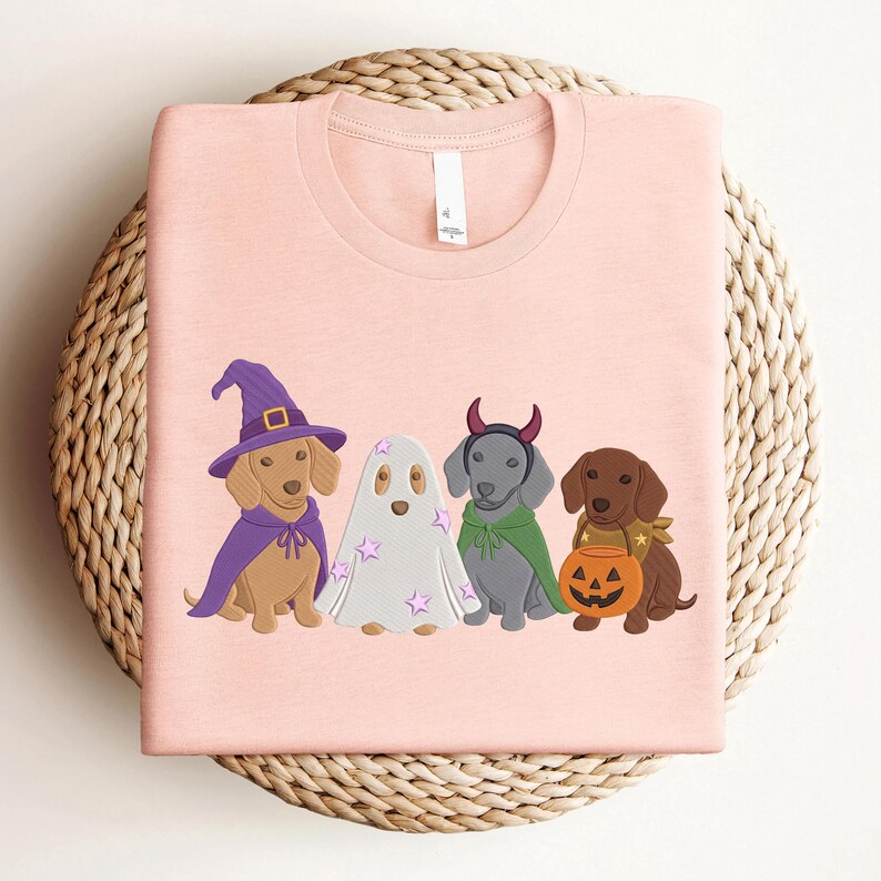 Puede incluir: Una camiseta rosa claro con un gr&aacute;fico de cuatro perros salchicha con disfraces de Halloween. Uno lleva un sombrero de bruja y capa morada, otro es un fantasma con estrellas rosas, otro tiene cuernos de diablo y capa verde, y otro una calabaza.