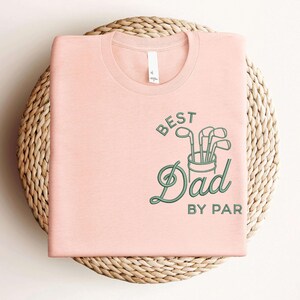May include: A light pink t-shirt with the text "BEST Dad BY PAR" in green, along with a graphic of golf clubs in a bucket. The shirt is folded and placed on a woven, circular surface.