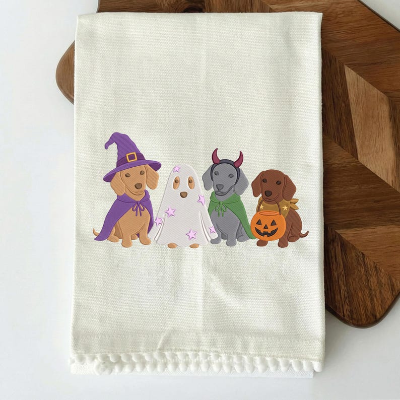 Puede incluir: Un pa&ntilde;o de cocina color crema con borde de pompones que presenta un gr&aacute;fico de cuatro perros salchicha con disfraces de Halloween: uno de bruja, uno de fantasma con estrellas rosas, uno de diablo con capa verde y otro con un cubo de calabaza.