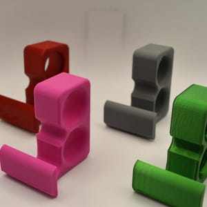 May include: Four brightly colored, 3D-printed phone stands. The stands are in red, pink, gray, and green. Each stand has a rectangular base and a vertical section with two circular openings. The stands are designed to hold a phone.