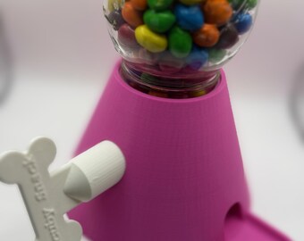 Op de afbeelding: Een roze snoepautomaat met een glazen pot vol kleurrijke snoepjes. De dispenser heeft een witte hendel met de tekst "Candy Snack". De dispenser is ontworpen om snoepjes te doseren.
