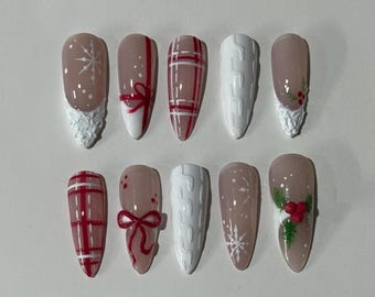 May include: A set of ten artificial nails with a winter holiday theme. Designs include snowflakes, red bows, plaid patterns, and white sweater-like textures. The nails are almond-shaped with a neutral base color and various festive accents.