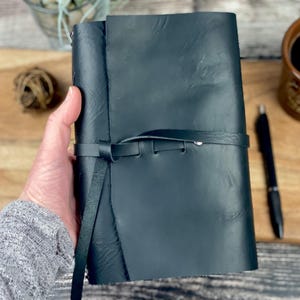 May include: A black leather journal with a wrap-around tie closure. The journal is closed and held in a hand. A black pen and a mug of coffee are on a wooden surface.