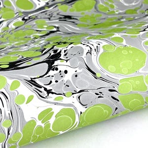 May include: A sheet of paper featuring a marbled pattern with vibrant lime green circular spots, interspersed with flowing streaks of black and shades of gray. The design creates a fluid, organic appearance across the surface of the paper.