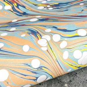 May include: A sheet of paper featuring a marbled pattern with swirling waves of light blue, peach, dark blue, and yellow. The design is accented by numerous solid white circular spots of varying sizes distributed across the colorful surface.