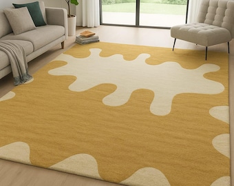 May include: Mustard yellow area rug with a large, off-white, abstract design. The rug is rectangular and placed on a hardwood floor. The room has a sofa, chair, and large windows.