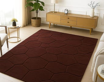 May include: A rectangular, burgundy area rug with a geometric pattern of connected, irregular hexagons. The rug is placed on a light-colored wooden floor in a living room setting. A mid-century modern style wooden cabinet and a potted plant are in the background.