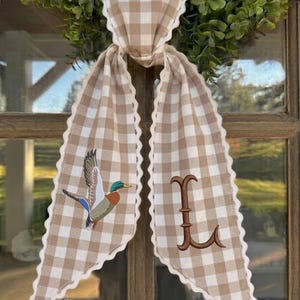 May include: A beige and white gingham fabric ribbon with scalloped edges, tied in a knot. The left tail features an embroidered mallard duck in flight, while the right tail displays a large, brown, embroidered letter L in a serif font.