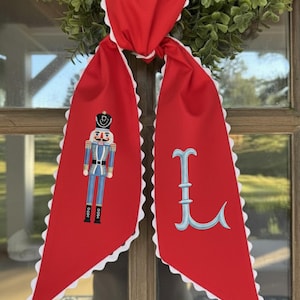 Puede incluir: Bufanda roja con borde festoneado blanco y nudo en la parte superior. Un lado presenta un soldado cascanueces en azul y dorado, el otro una letra "L" azul claro.