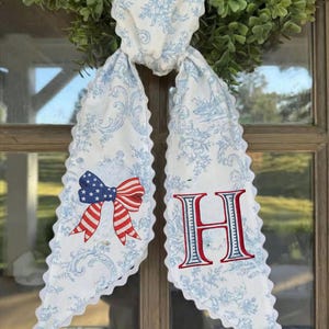 May include: A white and blue floral patterned door swag with a scalloped edge. One side features a red, white, and blue bow, the other a red and blue embroidered letter "H". The swag is tied to a green wreath.