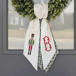 Puede incluir: Una cinta blanca para corona con un diseño de soldado cascanueces verde y rojo y una letra "B" roja. La cinta está drapeada sobre una corona verde, para decoración festiva.