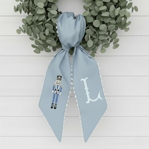 Puede incluir: Una bufanda de satén azul claro con un borde festoneado blanco y un diseño decorativo de cascanueces. La bufanda también presenta una letra "L" bordada en blanco. La bufanda está anudada y cuelga frente a una corona verde.