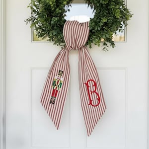 May include: A red and white striped ribbon with a green wreath in the background. The ribbon features a nutcracker design and a red letter "B" embroidered on it. The ribbon is tied in a bow.