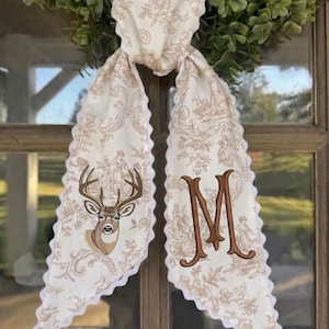 May include: A long, cream-colored fabric scarf featuring a tan toile floral pattern and a white scalloped edge. One end displays an embroidered brown deer head, while the other end features a large, embroidered brown letter M monogram.