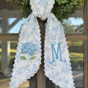 Könnte beinhalten: Ein weiß-blaues Türschild mit floralem Muster und gewelltem Rand. Das Türschild zeigt eine blau gestickte Hortensie und den Buchstaben "M". Das Türschild ist an einem grünen Kranz befestigt.