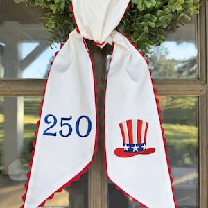 May include: White fabric door hanger with red scalloped edges and a green wreath. The hanger features the number "250" in blue embroidery and a patriotic Uncle Sam hat design.