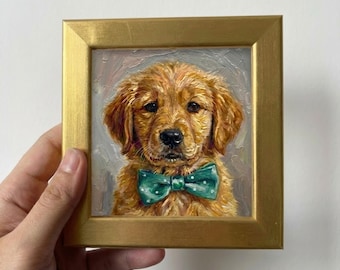 May include: A framed oil painting of a Golden Retriever puppy wearing a green bow tie with white polka dots. The painting has a gold frame and is set against a neutral background. The puppy has golden fur and a sweet expression.