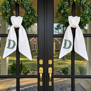 May include: Two green boxwood wreaths hanging on black double doors, each adorned with a white fabric ribbon featuring a scalloped edge and a dark green embroidered letter D monogram centred on the lower portion of the hanging fabric tails.