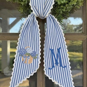 Op de afbeelding: Een blauw-wit gestreepte stoffen deurhanger met een geschulpte rand. De deurhanger heeft een bloemenmandontwerp en een blauw geborduurde letter "M". Het is bevestigd aan een groene krans.