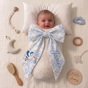 May include: A baby wrapped in a white blanket with a large blue and white bow. The bow features a stork carrying a baby and the name "James" embroidered in blue. Accessories include a pacifier, wooden toys, and a baby brush.