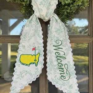 May include: White and green fabric door hanger with a floral pattern and scalloped edges. Features an embroidered yellow and green golf course map with a red flag, and the word "Welcome" in green script.
