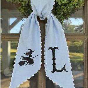 May include: A light blue fabric door hanger with scalloped edges. The left side features a black embroidered silhouette of a witch flying on a broomstick, while the right side displays a large, black embroidered capital letter L in a serif font.