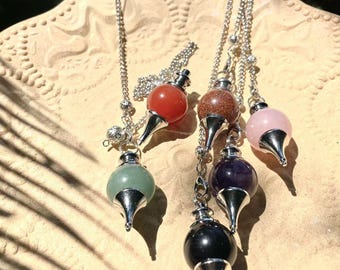 May include: A collection of crystal pendulums with silver chains. The pendulums feature various colored gemstones, including red, brown, pink, purple, green, and black. Each pendulum has a silver-colored metal tip and a round gemstone at the top.