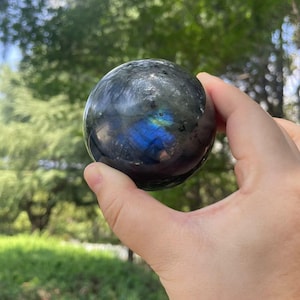 以下が含まれることがあります： 研磨された球形のラブラドライトの宝石。球体は主に濃い灰色で、虹色の青と緑のきらめきがあります。ぼやけた緑の葉の背景に対して、石は手で持たれています。