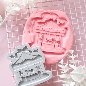 Puede incluir: Un cortador de galletas gris claro con forma de carrusel y una impresión de arcilla rosa a juego que presenta una atracción de feria detallada con caballos, un toldo y un lazo en la parte superior. El diseño muestra un carrusel clásico.