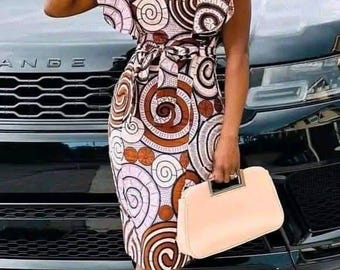 Pode incluir: Vestido estampado com decote em V e mangas com babados. O vestido apresenta um design espiral marrom, branco e laranja. O traje é completado com um chapéu a condizer, óculos de sol, uma bolsa clara e saltos.