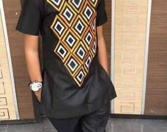 Puede incluir: Una persona joven vestida con una camisa negra de manga corta con un diseño geométrico en dorado, blanco y negro. El conjunto incluye pantalones negros, zapatos rojos y gafas de sol. También se ve un reloj de pulsera.