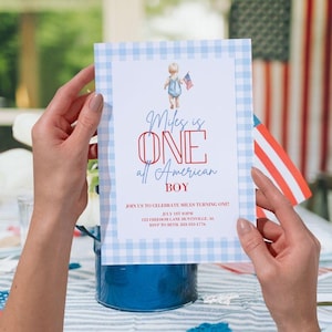 May include: A blue and white gingham patterned invitation with the text "Miles is ONE all American BOY" is held by a person. The invitation features a watercolor illustration of a child holding a small American flag. The invitation is for a first birthday.