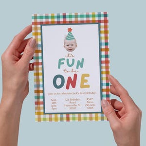 May include: A colorful birthday invitation with a plaid border is held by two hands. The invitation features the text "It's FUN to be ONE" with a baby's photo and event details. The invitation is for a first birthday.
