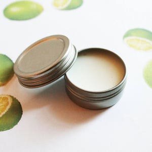 May include: An open, small, round, silver-colored tin with a white, creamy substance inside. The lid is next to the tin. The background features lime illustrations in various sizes and orientations, with a white background.