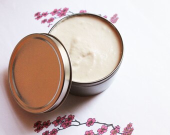 May include: An open, round, silver-colored tin with a light beige cream inside. The lid is next to the tin, showing a matching silver rim and a light brown interior. The background is white with pink floral accents.