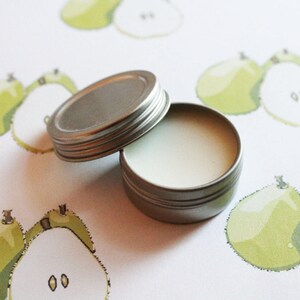 May include: A small, open, silver-colored tin of balm sits on a white surface decorated with green and yellow pear illustrations. The balm is white and the tin's lid is next to it.